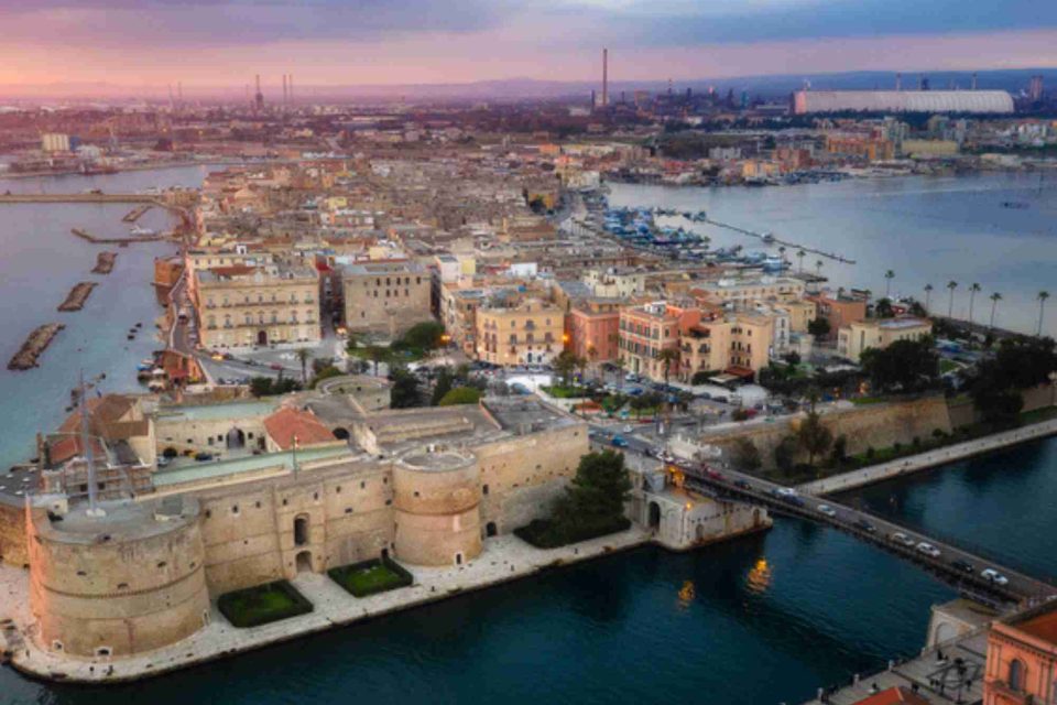 Veduta aerea di Taranto con il Castello Aragonese in primo piano e la città sullo sfondo. Si vedono il Ponte Girevole e le acque del mare che circondano la città, con un cielonon limpido come dovrebbe